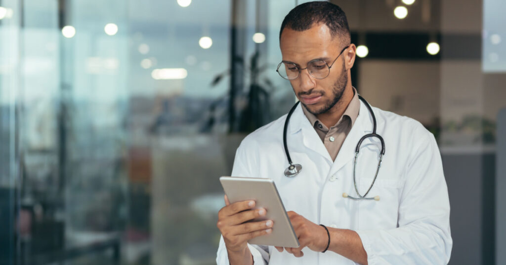 Serious and focused thinking african american doctor using tablet computer, man working inside modern clinic office.