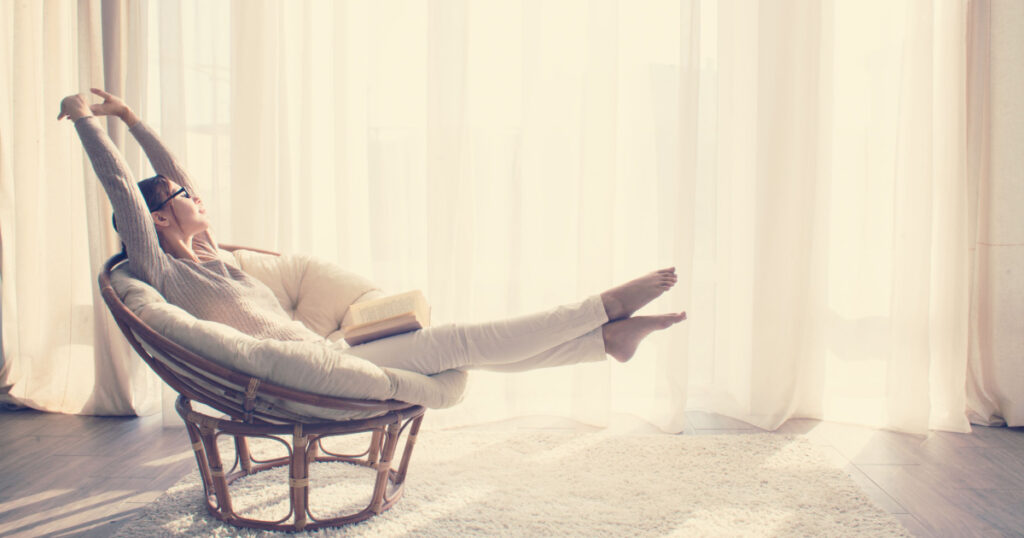 Young woman at home sitting on modern chair in front of window relaxing in her living room reading book, instagram toning