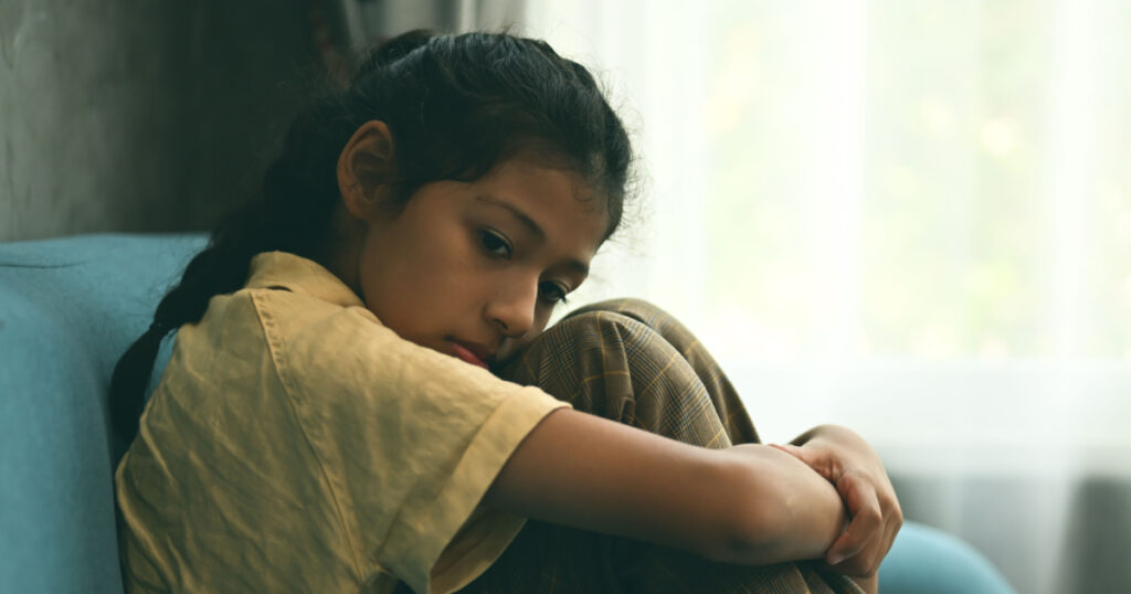 Unhappy girl portrait, Sad child sitting on sofa at home, Upset girl hugging knees alone, Concept of lonely girl and kid with trouble and violence