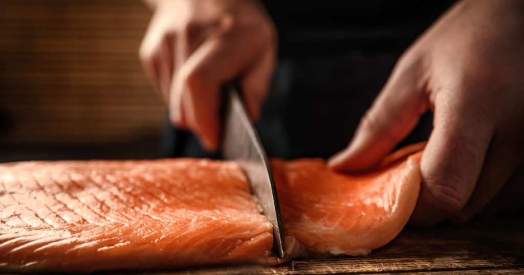Chief hands cut salmon fillet with knife on wooden table at kitchen. Man cooking red omega fish with lemons for healthy nutrition diet