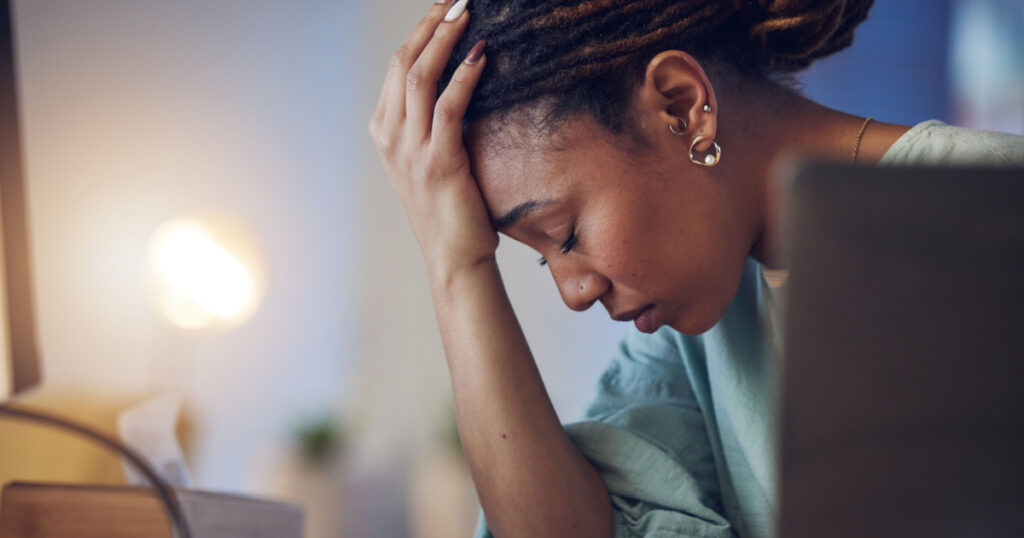 Business woman, depression and stress in an office at night working late on deadline. Tired African entrepreneur person with hands on head for pain, burnout or regret for mistake or fail at work
