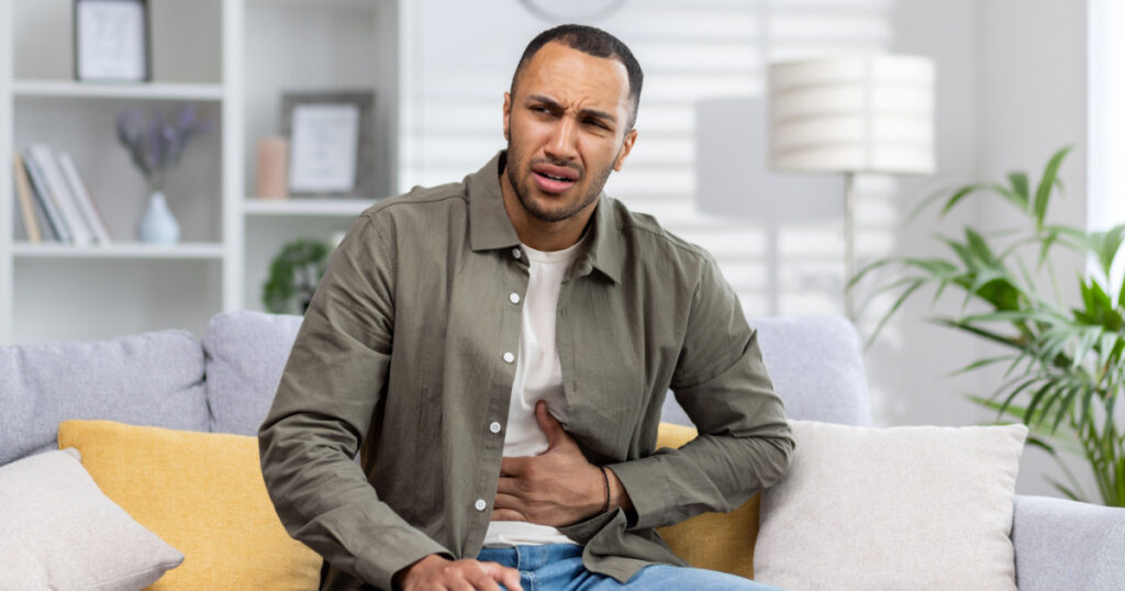 A young African American man is suffering from abdominal pain, stomach cramps and constipation. He is sitting on the sofa and holding his stomach, grimacing in pain.