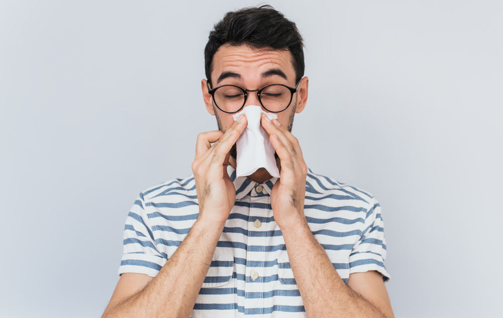 Horizontal portrait of unhealthy handsome man wearing striped shirt and glasses, blowing nose into tissue. Male have flu, virus or allergy against white background. Healthy medicine and people concept
