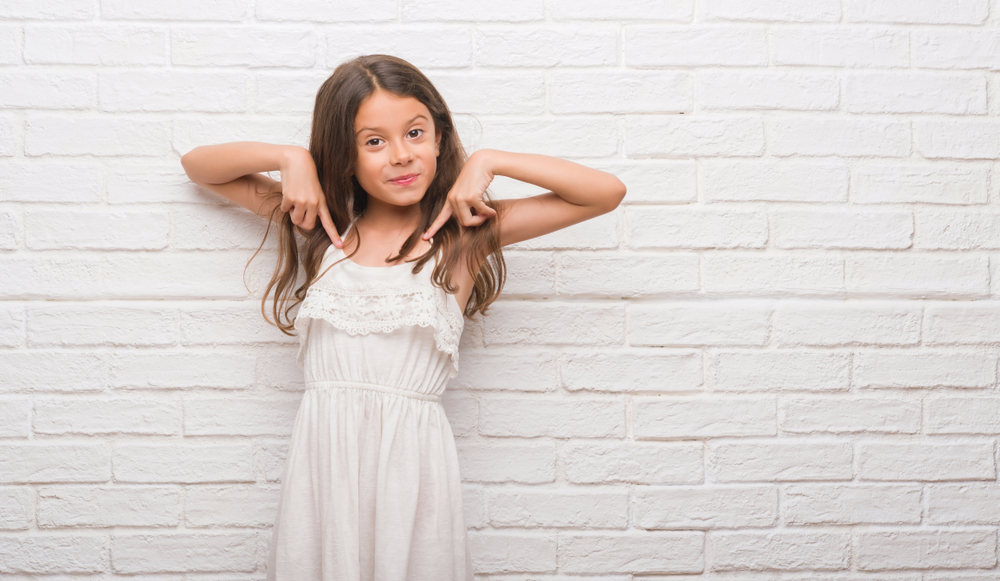 Young hispanic kid over white brick wall looking confident with smile on face, pointing oneself with fingers proud and happy.