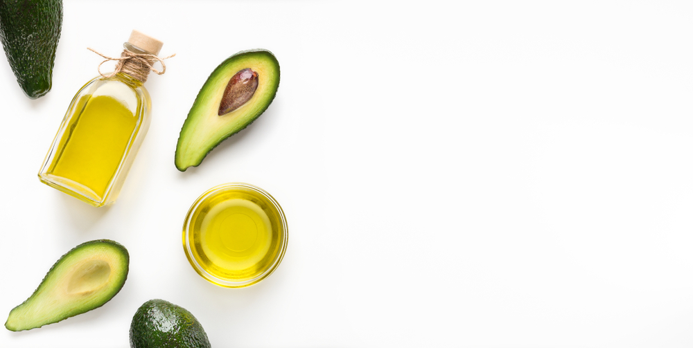 Sliced avocadoes and oil in bowl and bottle on white background top view, copy space. Vegetarian oils concept