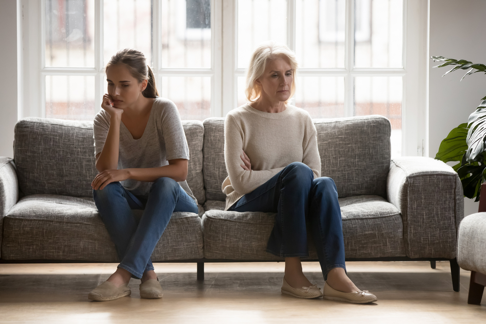 Frustrated upset young grown up daughter and middle aged retired mother sitting separately in silence on sofa at home, stressed after quarrel, ignoring each other, feeling depressed, generation gap.
