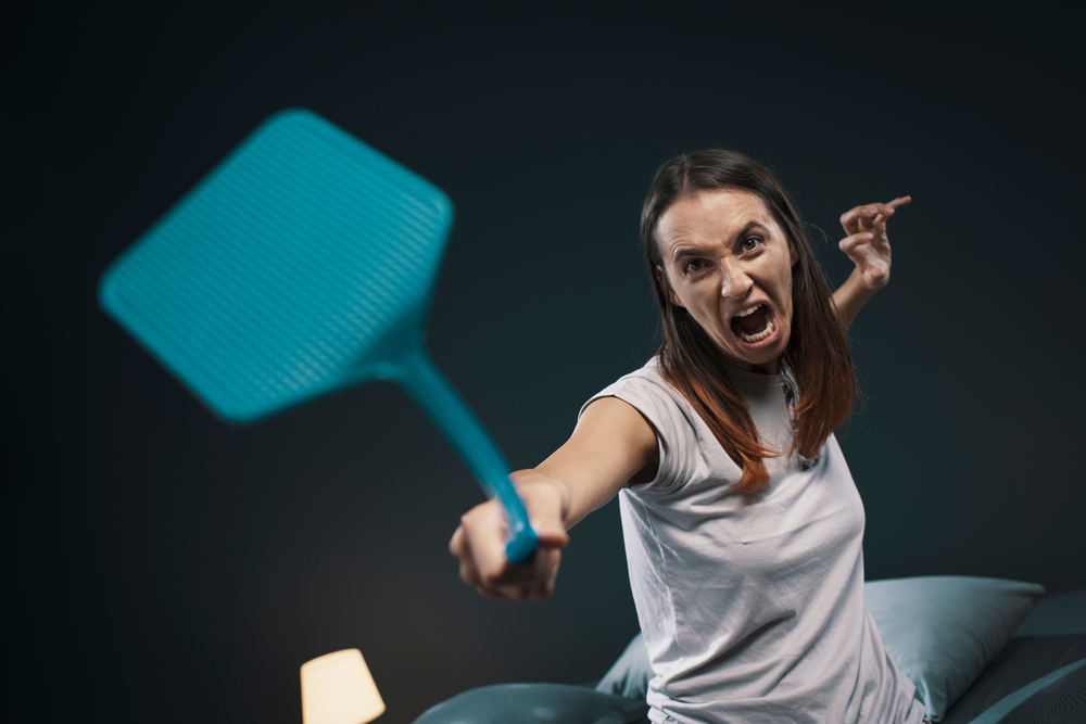 Angry woman killing mosquitoes in her bedroom, she is sitting on bed and holding a fly swatter like a weapon