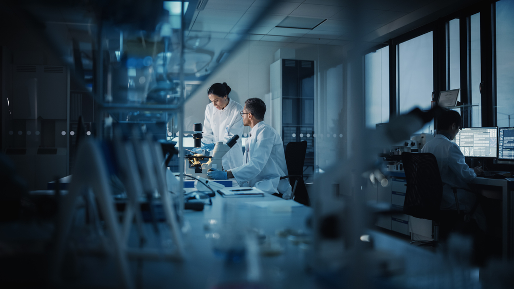 Modern Medical Research Laboratory: Two Scientists Working, Using Digital Tablet, Analyzing Test, Talking. Advanced Scientific Pharmaceutical Lab for Medicine, Biotechnology Development. Evening Time
