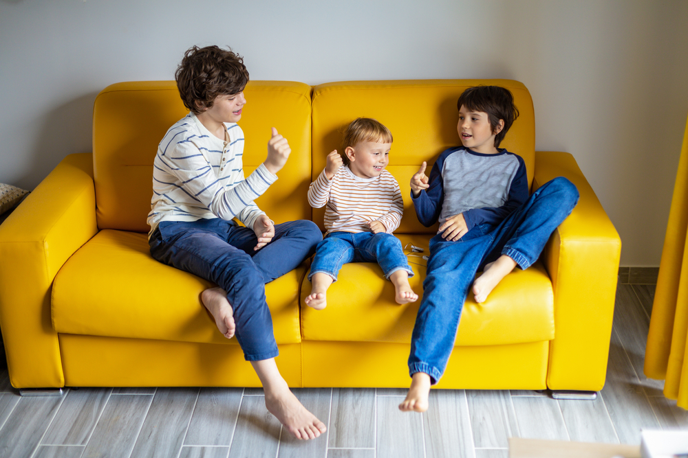 Three brothers play rock, paper, scissors game together sitting on yellow sofa at home. Siblings love spend time together during holidays.