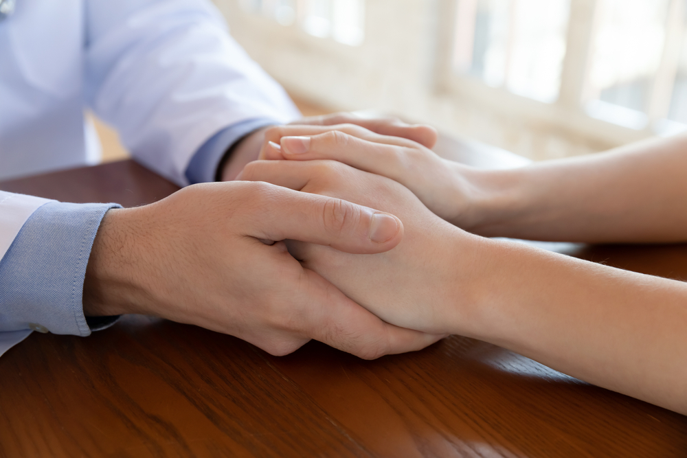 Close up compassionate male gp doctor holding hands of stressed young female patient, giving psychological help, supporting soothing after getting bad health test results at checkup meeting.