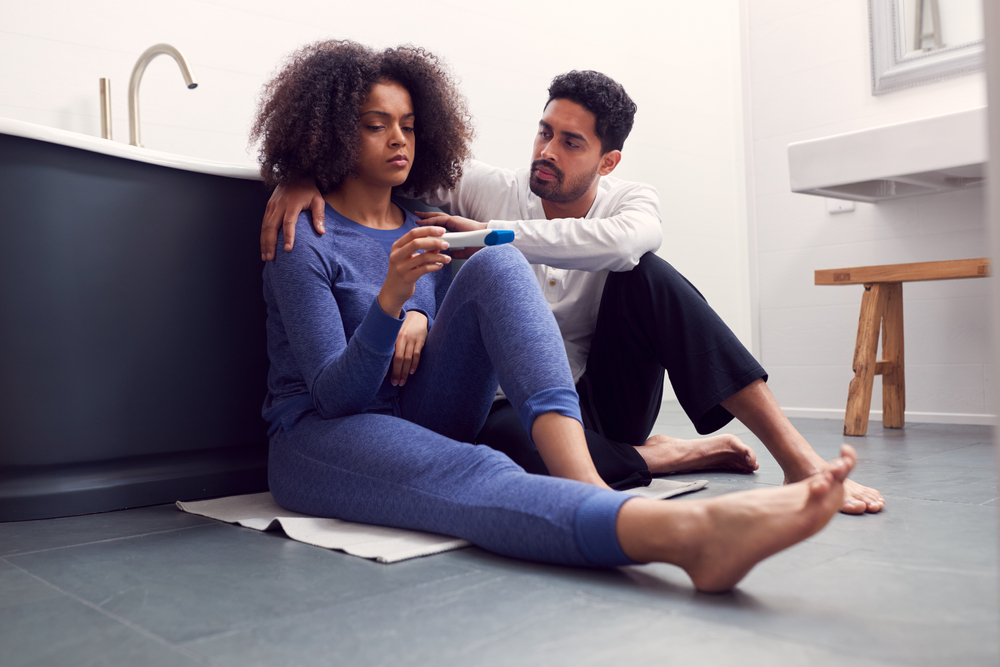 Disappointed Couple Sitting On Floor In Bathroom At Home With Negative Home Pregnancy Test
