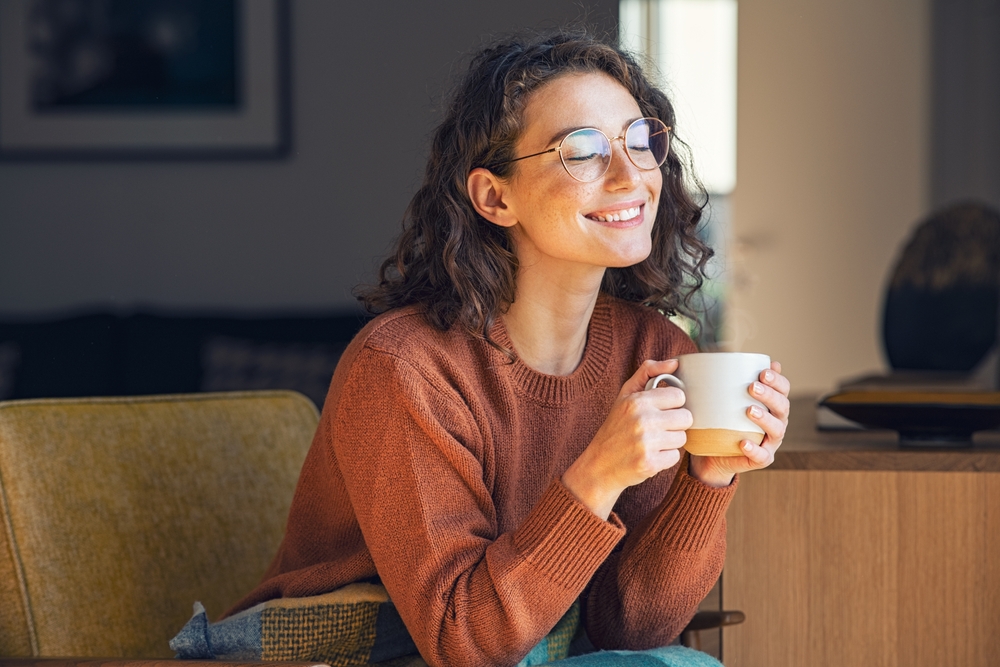 Happy young woman drinking a cup of tea in an autumn morning. Dreaming girl sitting in living room with cup of hot coffee enjoying under blanket with closed eyes. Pretty woman wearing sweater at home.
