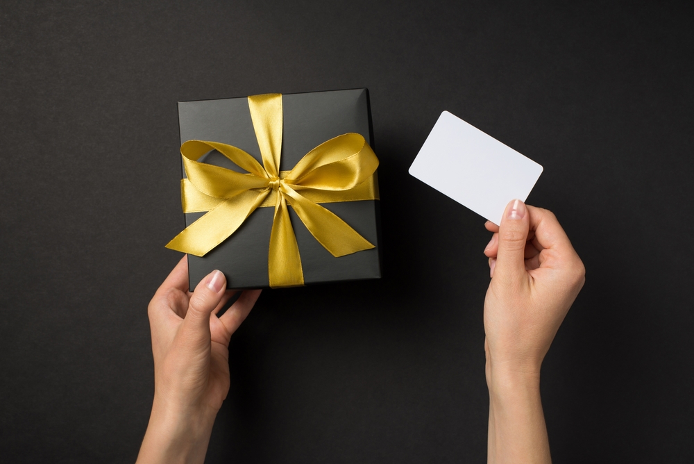 First person top view photo of hands holding black giftbox with golden satin ribbon bow and white plastic card on isolated black background with blank space