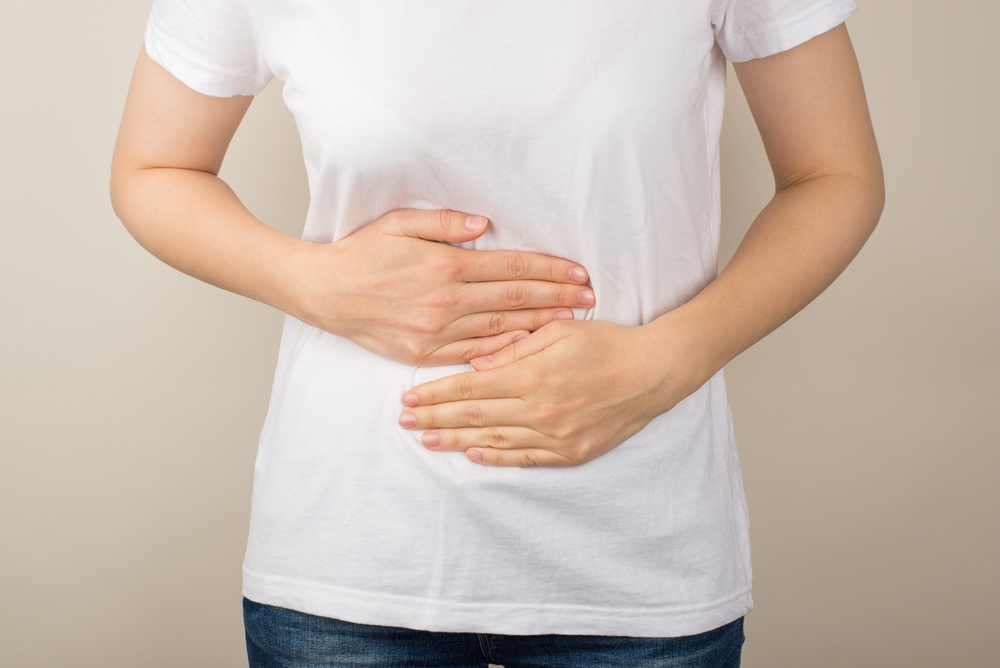 Cropped closeup photo of young woman in white t-shirt and jeans holding her hands on upper abdomen on isolated grey background