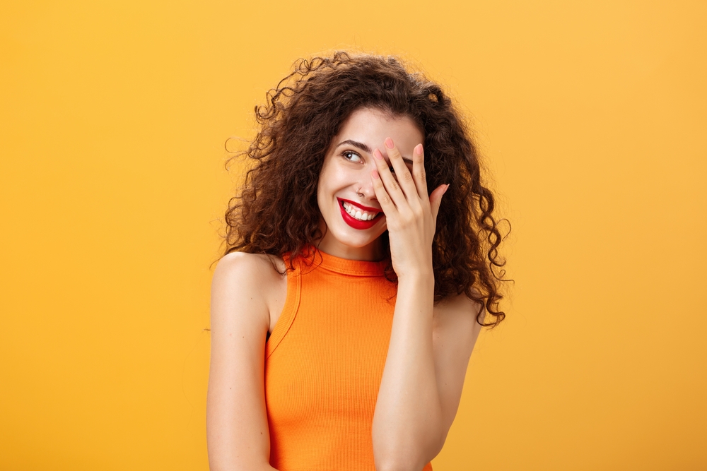 Woman being praised by coworkers feeling awkward and shy hearing congratulations with received award covering face peeking left with joyful satisfied smile posing over orange background
