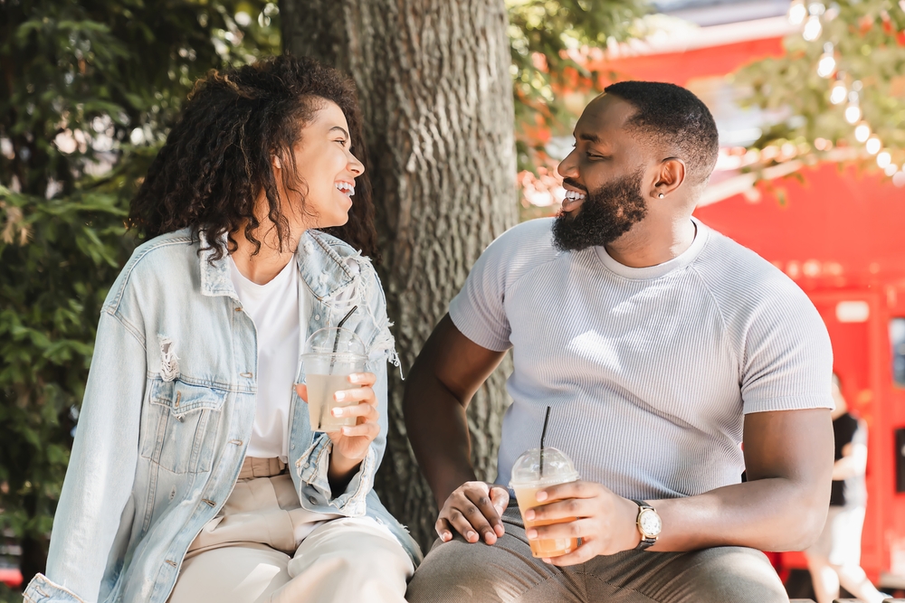 African-american romantic couple having romantic date, drinking lemonade while sitting talking communicating in park outdoors. Love and relationship, bonding concept

