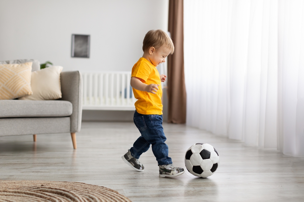Future champion. Adorable little toddler boy playing football, hitting ball at home, having fun in living room interior, copy space