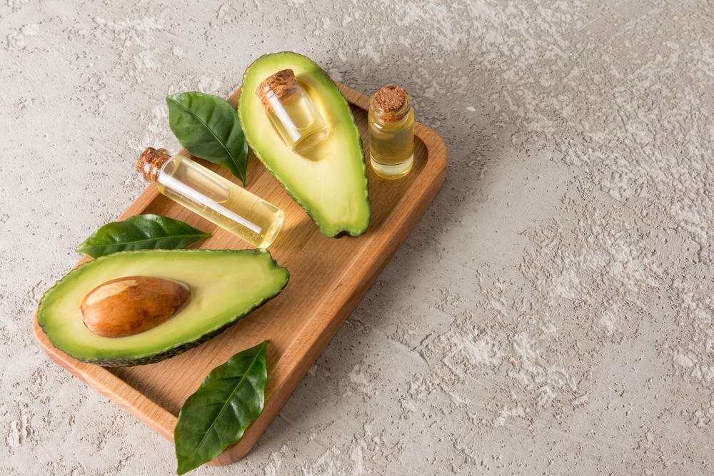 Bowl with oil, fresh cut avocados and leaves on light green background, flat lay. Space for text