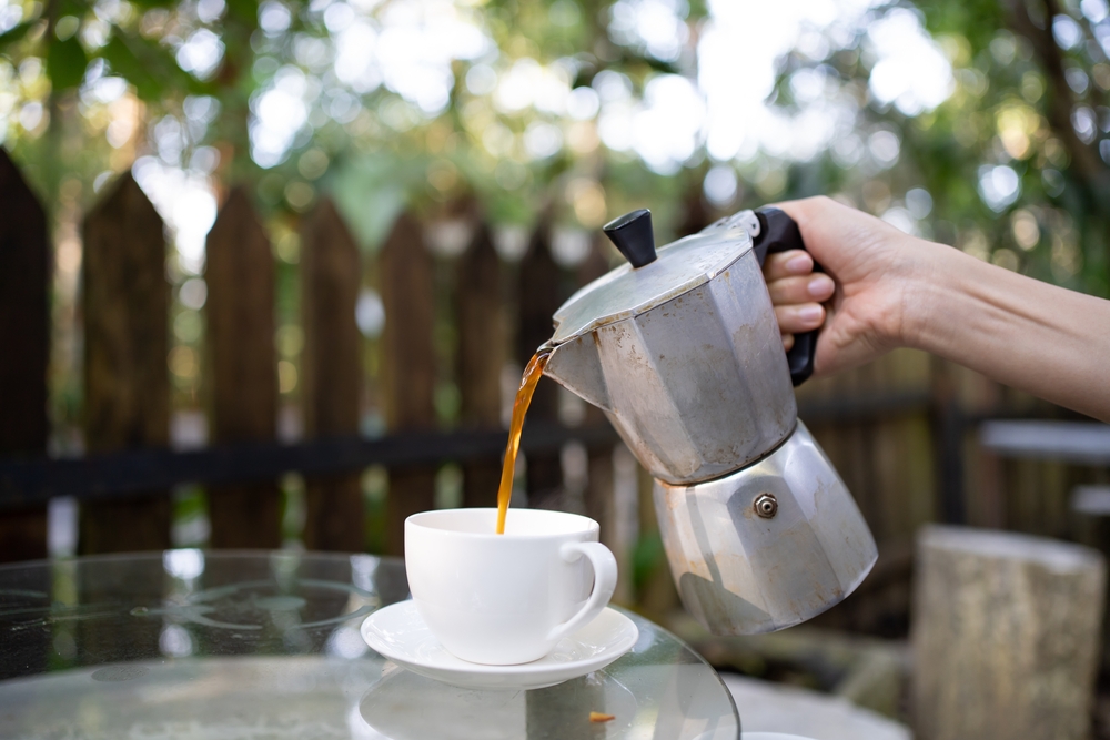 Lady hand pouring freshly brew coffee with Moka Pot with blur country side forest background. Morning brew coffee. Italian classic vintage coffee maker
