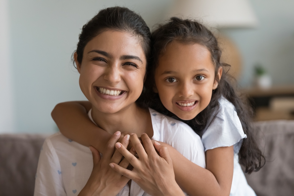 Happy beautiful young Indian mom and cute daughter kid home head shot. Cheerful mother piggybacking child looking at camera with toothy smile, hugging, talking on video call. Screen view portrait
