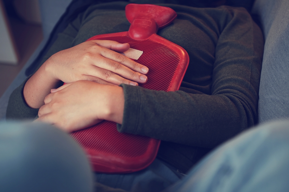 Asian woman has sick heavy menstrual pain applying red hot water bag on her stomach to relieve the pain on the living room sofa