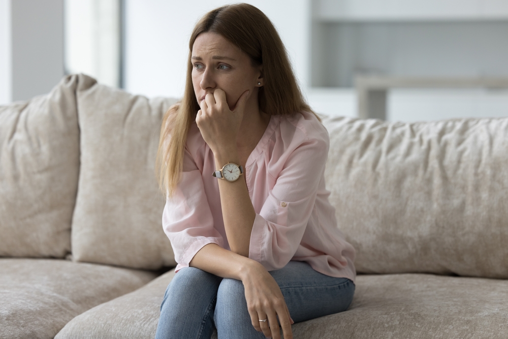 Unhappy depressed pretty woman sitting on sofa at home, looking away, thinking over health problems, crisis, loss, bad news, touching face, feeling stressed, frustrated. Negative emotions