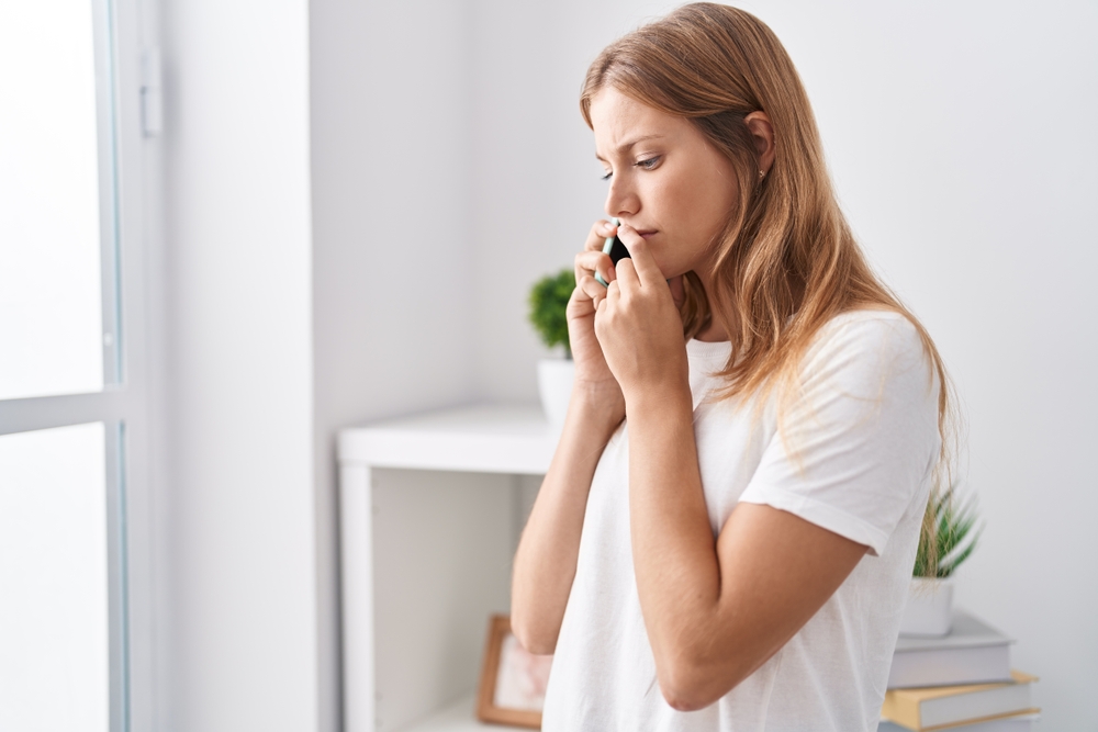 Young blonde girl talking on smartphone with worried expression at home