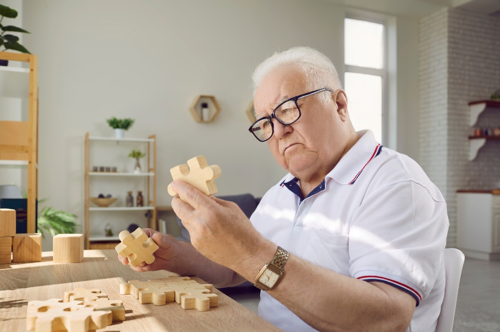 Senior man playing with jigsaw parts. Old man with Alzheimer's disease sitting at desk in geriatric clinic or nursing home, playing game, looking at wood puzzle piece he's holding in hand and thinking