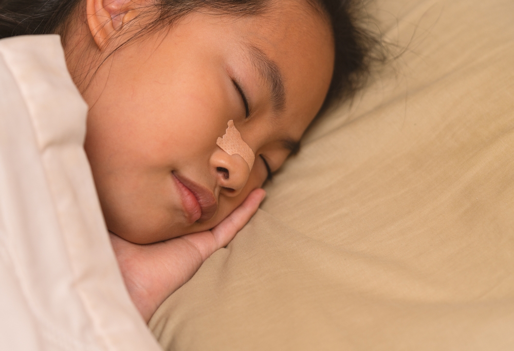 Asian child girl is sleeping well in bed, close up to the side of her face on pillow, nasal strip at her nose. Asian child girl at 8 or 9 years old. Blank space for copy and design.