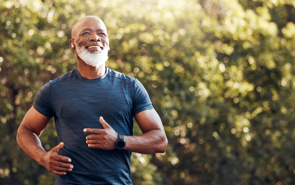 Happy black man running in park with music, smile and mockup in nature, garden and workout. Senior male, outdoor runner and motivation for fitness, energy and healthy exercise training with earphones
