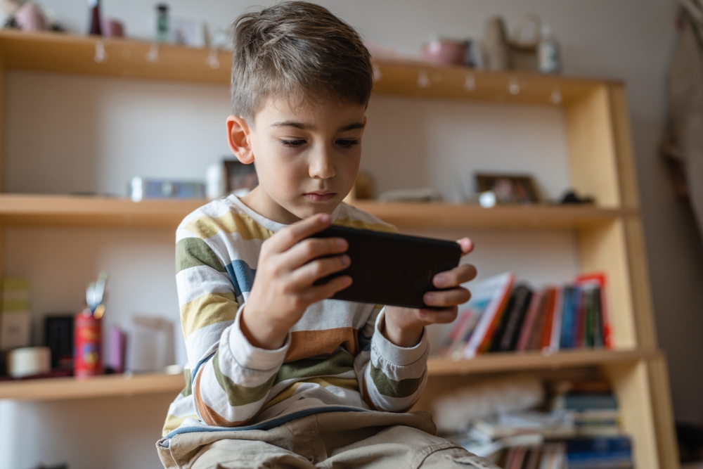 one boy caucasian child preschooler hold smartphone mobile phone at home play video games childhood and growing up technology addiction concept use smartphone app for online browsing or watch video
