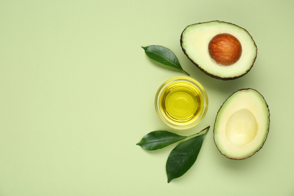 Bowl with oil, fresh cut avocados and leaves on light green background, flat lay. Space for text