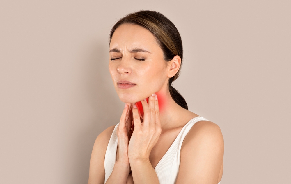Unhappy young brunette woman with closed eyes wearing white top suffering from pain in throat, touching red inflamed zone on her neck, panorama with empty space, beige studio background