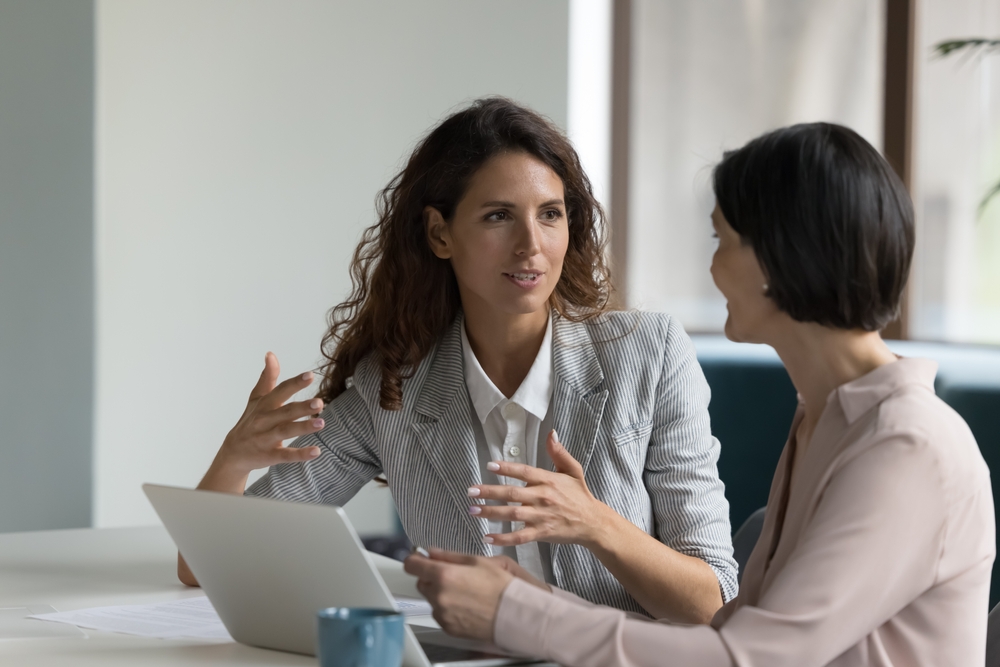 Two business women sit at desk discuss project details, diverse female colleagues met in office, share opinion, working on collaborative task, sales manager makes commercial offer to company client
