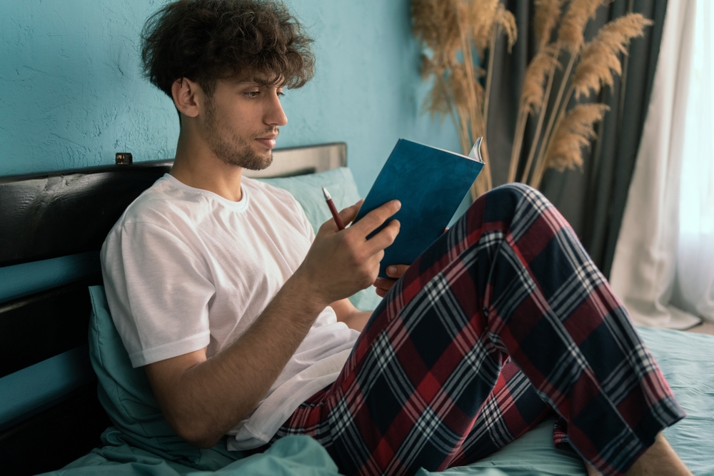 Man in pajamas sitting on his bed writing notes in journal. Copy space