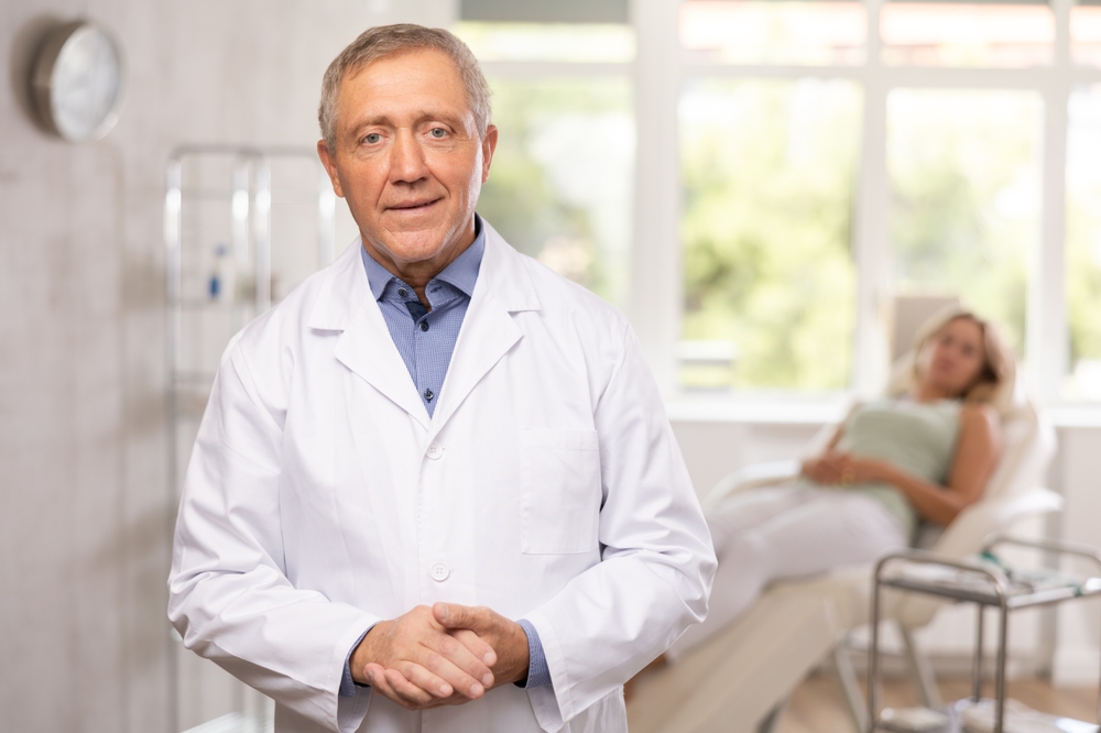 Positive old man doctor posing against background of doctor's cabinet with clinical chair
