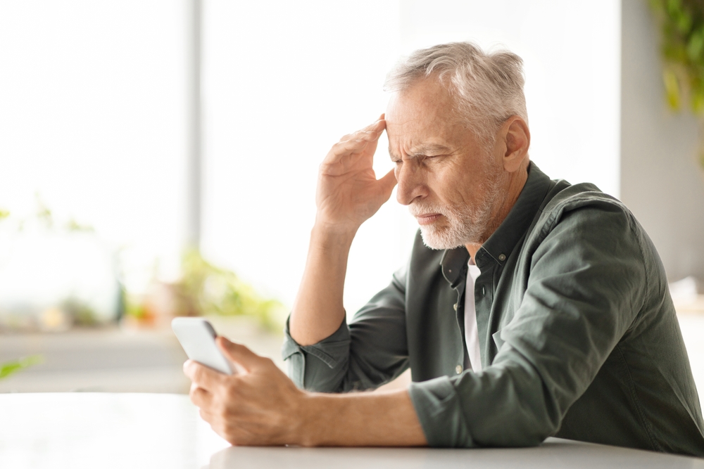 Distressed elderly man suffering headache while looking at smartphone at home, worried senior gentleman reading troubling news or navigating complex app, sitting at table in kitchen, copy space
