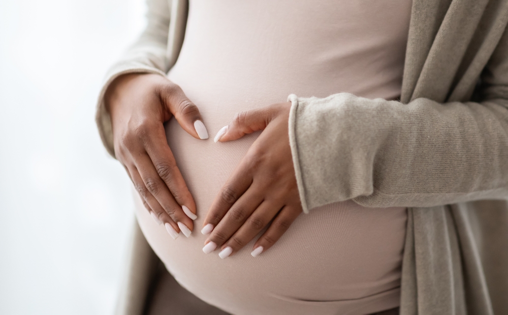 Pregnant black woman holding palms in heart shape on her big tummy, cropped. Unrecognizable african american expecting lady standing by window at home. Pregnancy and maternity concept