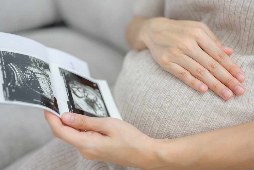 Close-up of a pregnant woman sitting in the living room at home looking at the ultrasound image of her own baby. Happy pregnancy woman during prenatal ultrasound at her home