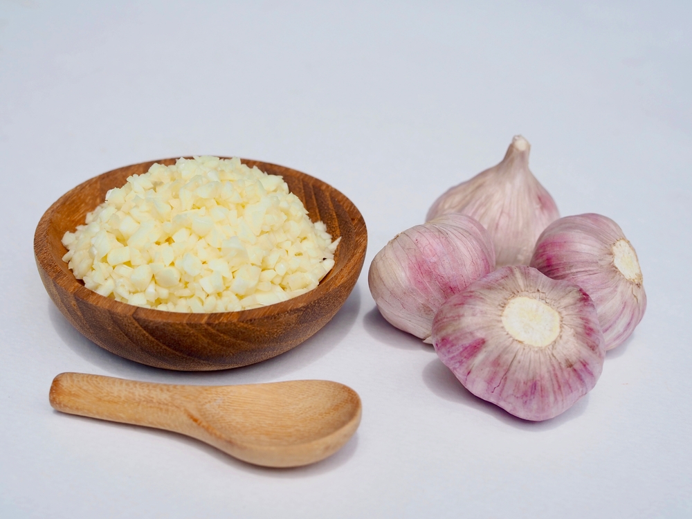 Chopped garlic in a wooden bowl and garlic and a wooden spoon are placed on a white background.
