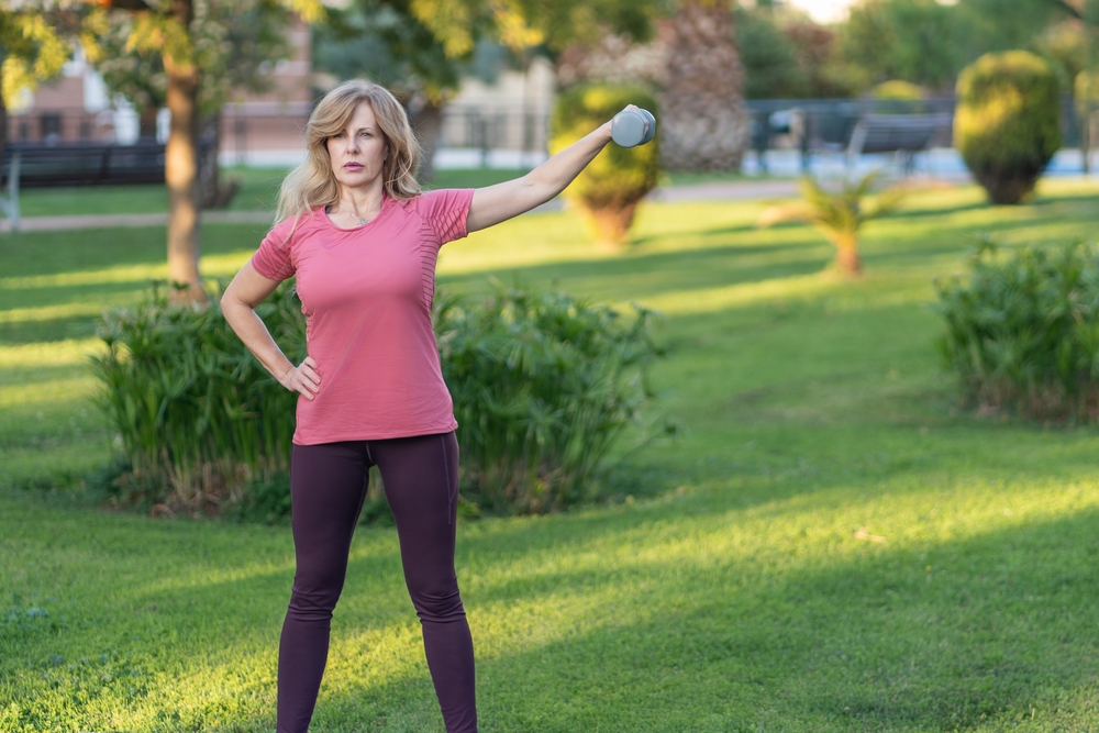 Woman performs lateral arm raise with weight in a sunny park setting.
