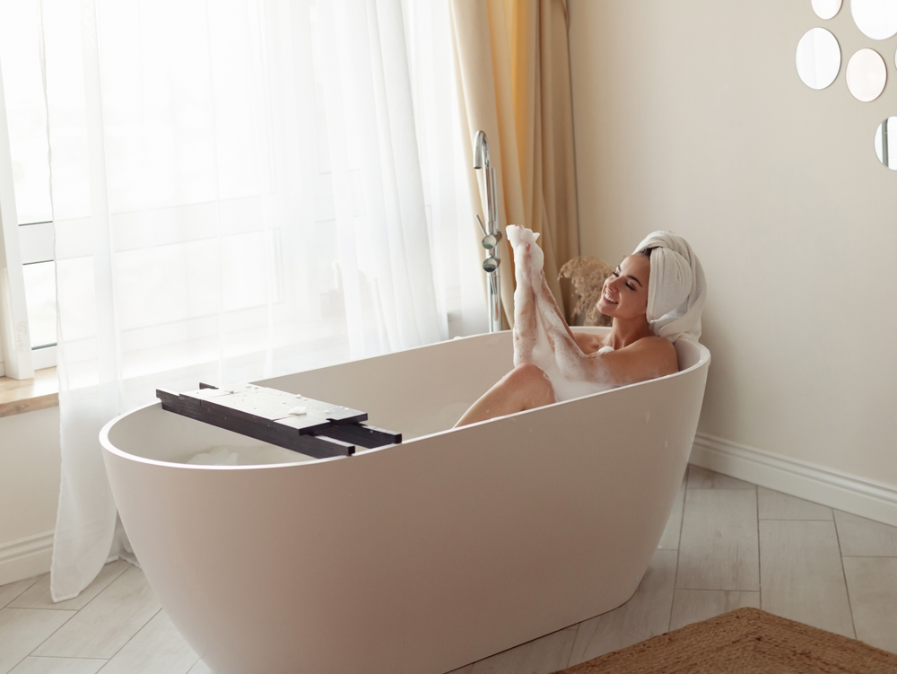 Cheerful young Caucasian beautiful positive female lying in bathtub with foam in good mood smiling and washing body relaxing in bathroom. Morning hygiene, body care routine. Bath relaxation
