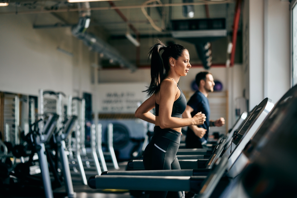 Side profile of a fit girl running on a treadmill at the gym.
