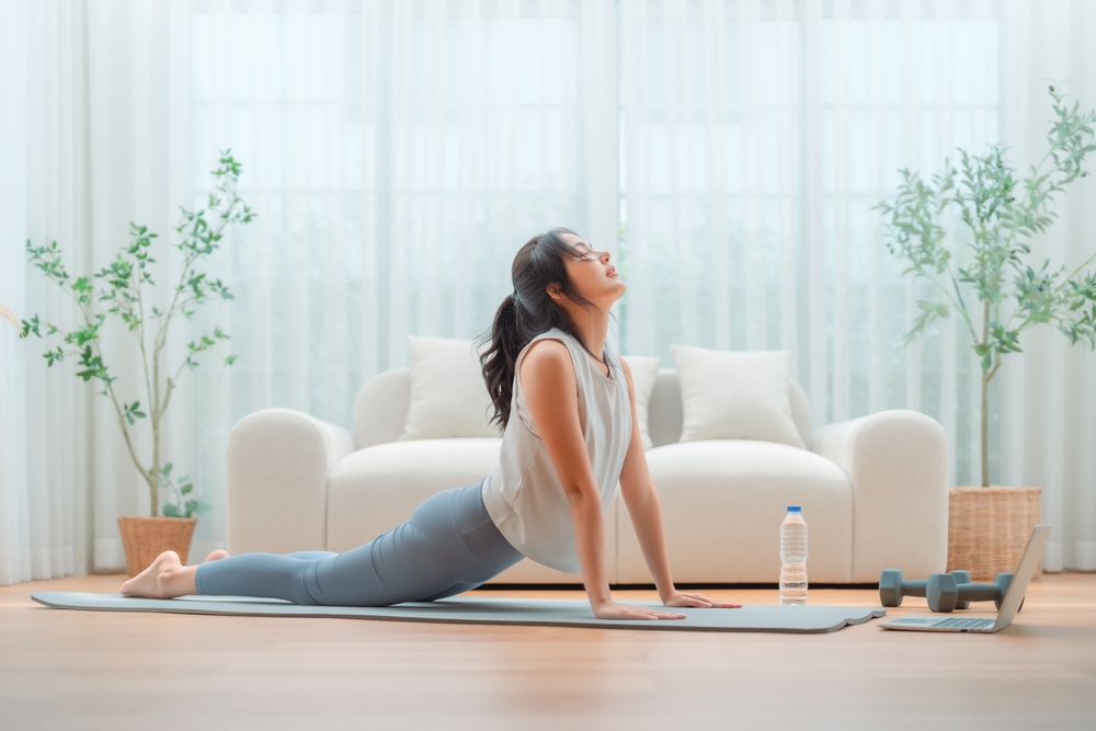 Young Asian woman stretching yoga workout on exercise mat while online training class with computer laptop in living room