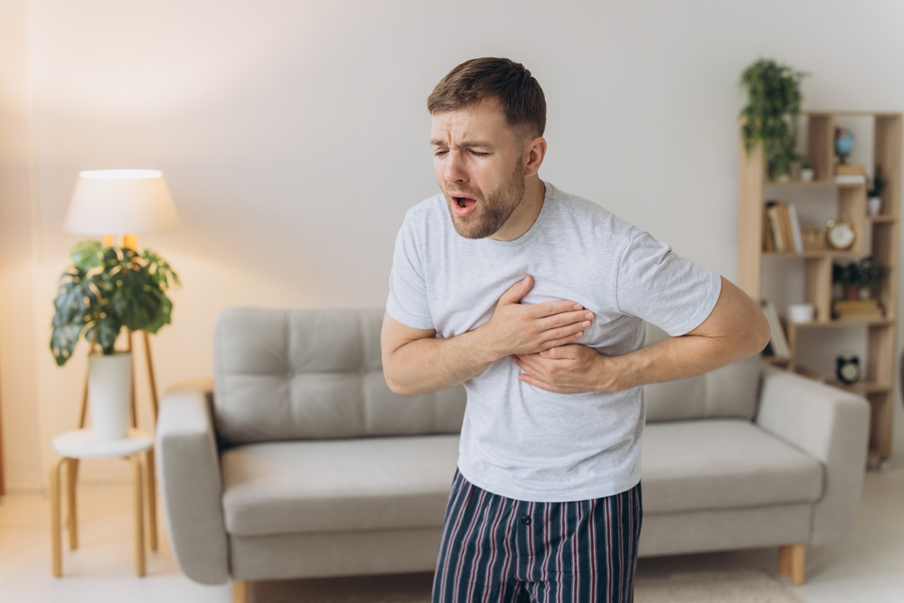 A young man is standing at home and holding his chest. Feels severe pain, shortness of breath, panic attack, heart attack, stroke.
