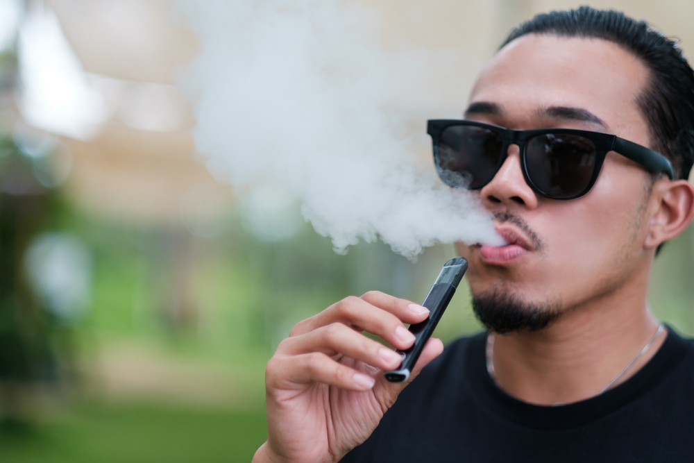 Asian man vaping with an electronic cigarette vape device concept. Smoking with an E-cigarette blowing smoke from mouth, and enjoying outdoors in summer, modern alternative to tobacco addictive habit.
