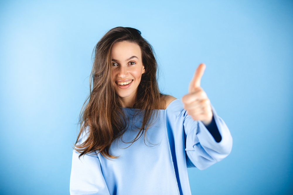 Optimistic brunette hair woman makes thumbs up gives approval shows excellent gesture says nice job gives positive feedback shares good opinion, wow, wear blue top isolated over blue background.
