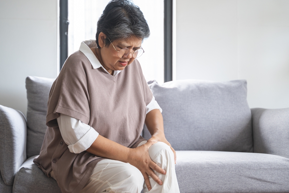 Body arthritis muscles stiff problem, aged woman suffering on leg, pain knee from disease chronic, hand massage rubbing joint injury, mature hurt or sore sitting on sofa at home. Health care people.
