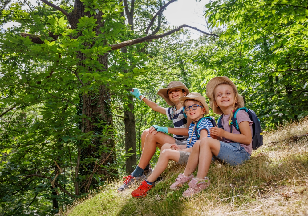 Three siblings sit on forest hill take a rest after trail hike with summer school in sunny woods