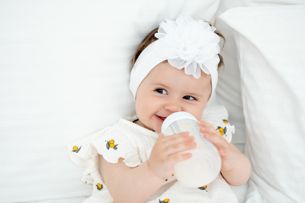 a baby with a bottle of milk or formula in his hands in a bright bedroom, a space for text, a baby food concept, a small child sucking a pacifier with a bottle.
